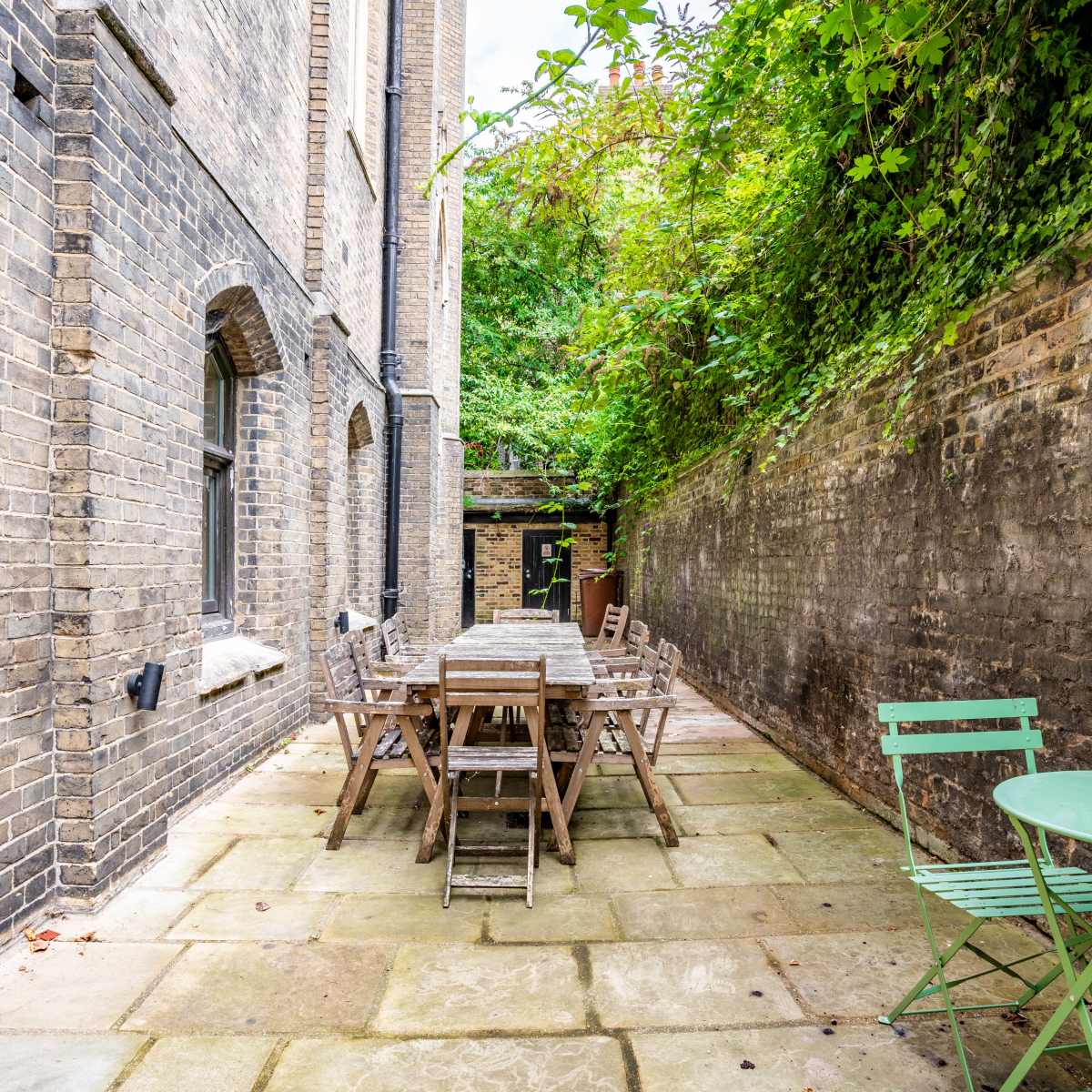 A narrow, paved courtyard features a long wooden dining table surrounded by chairs, flanked by brick walls and overgrown greenery.