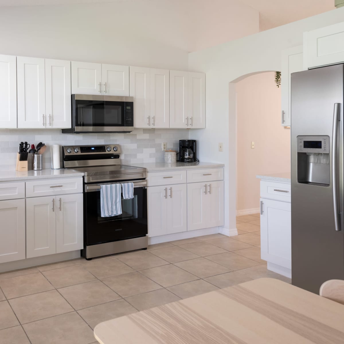 The kitchen features white cabinetry, stainless steel appliances, and a tiled backsplash, with a countertop and table visible in the foreground.