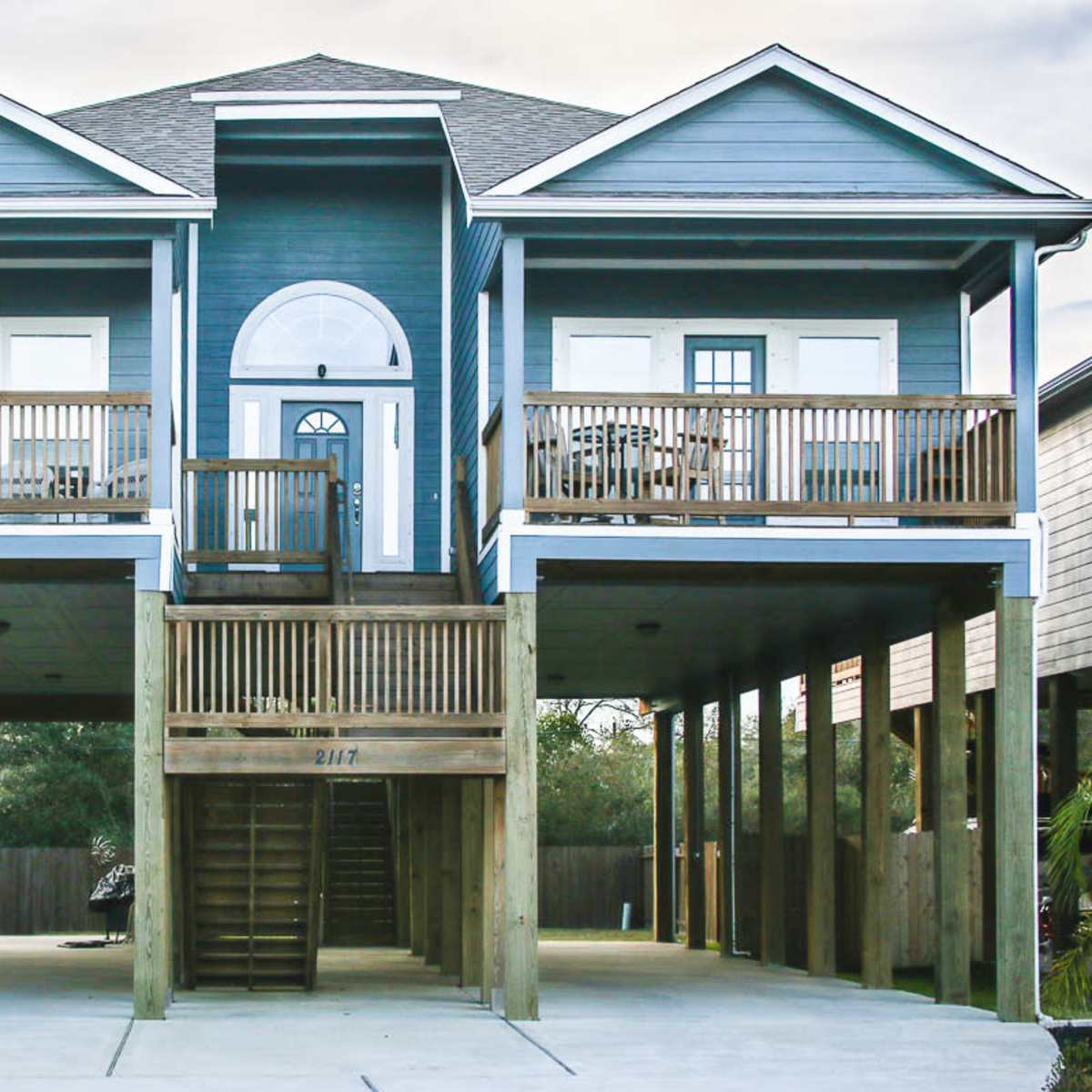 A two-story blue house with a central front entrance and balconies on both sides is supported by wooden stilts.