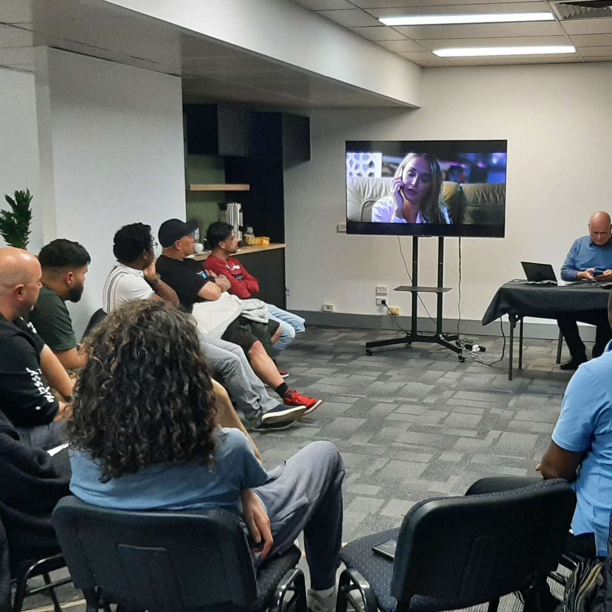 A group of people sits in a room, watching a video on a large screen while another person works at a table nearby.