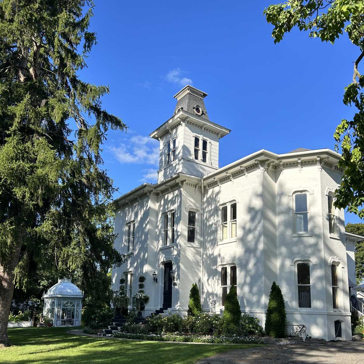 A large white historic house with a clock tower, surrounded by green grass and trees, and a small glass greenhouse nearby.