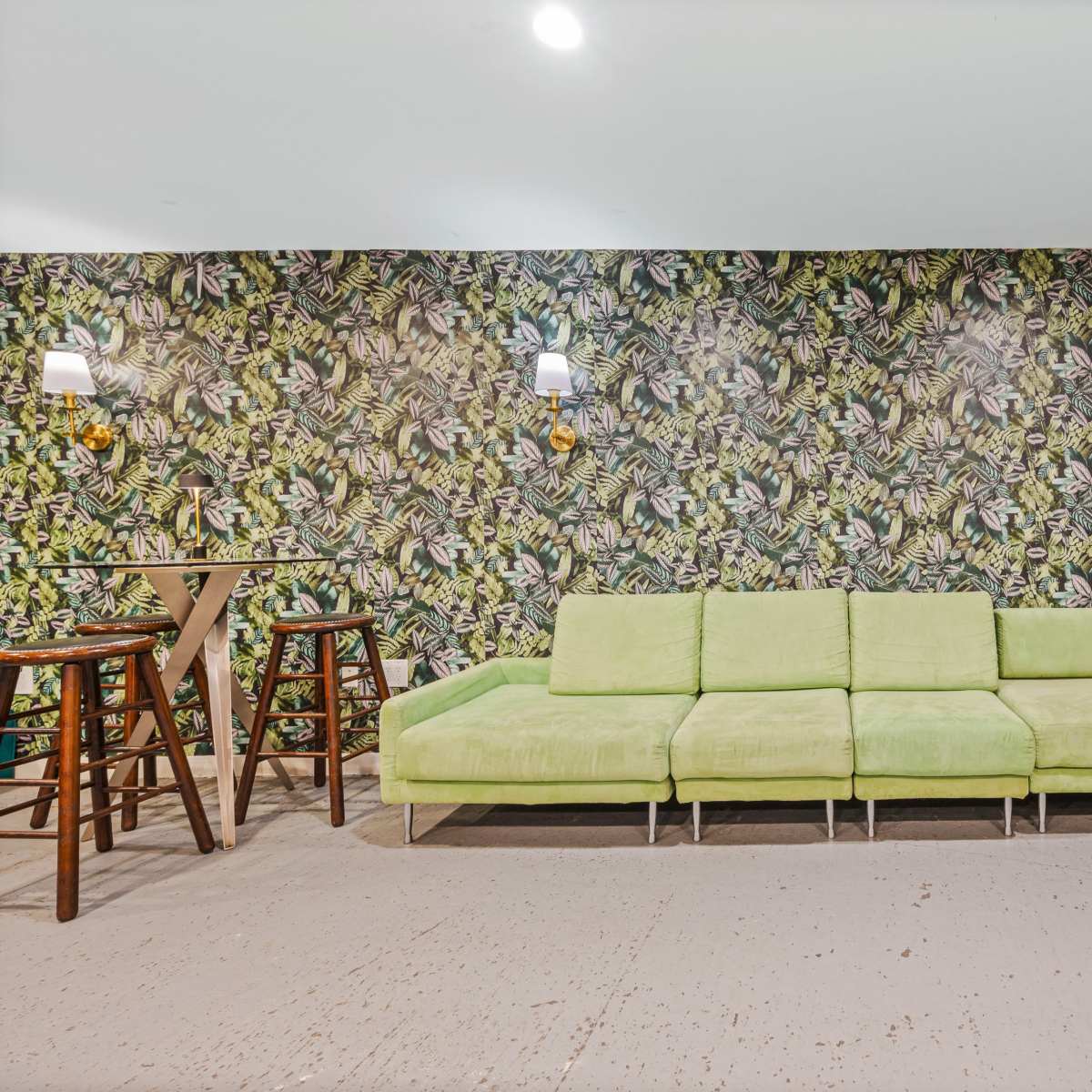 A green sectional sofa against a vibrant botanical-themed wallpaper, with wooden bar stools and a small table nearby.
