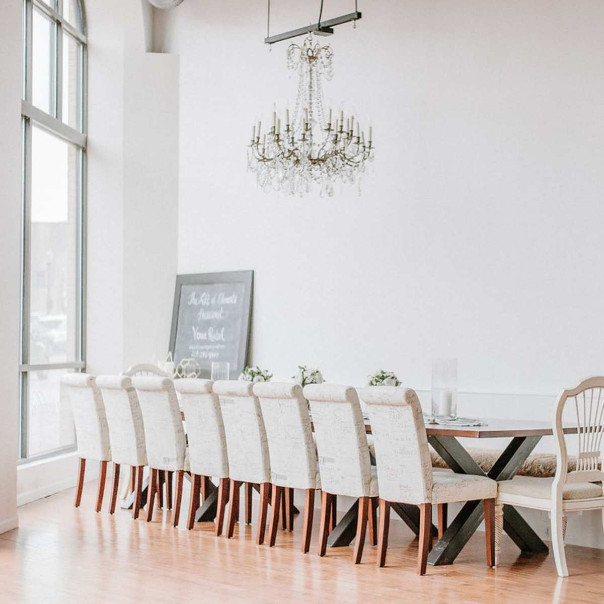 A long wooden dining table is set in a bright room with a chandelier above and a large chalkboard on the wall.