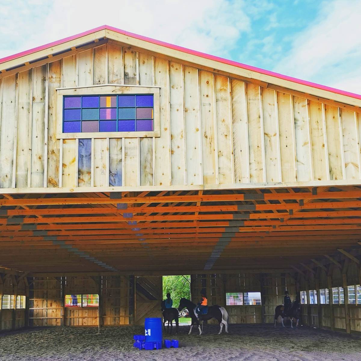 A wooden horse barn with a colorful stained glass window, where two horses are being ridden inside.