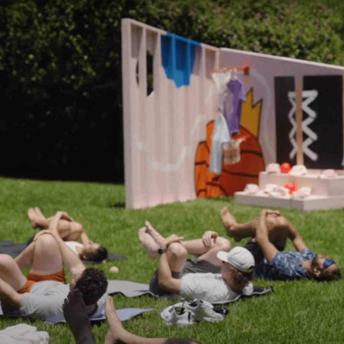 A group of people lies on the grass in front of a colorful backdrop displaying clothing and art.