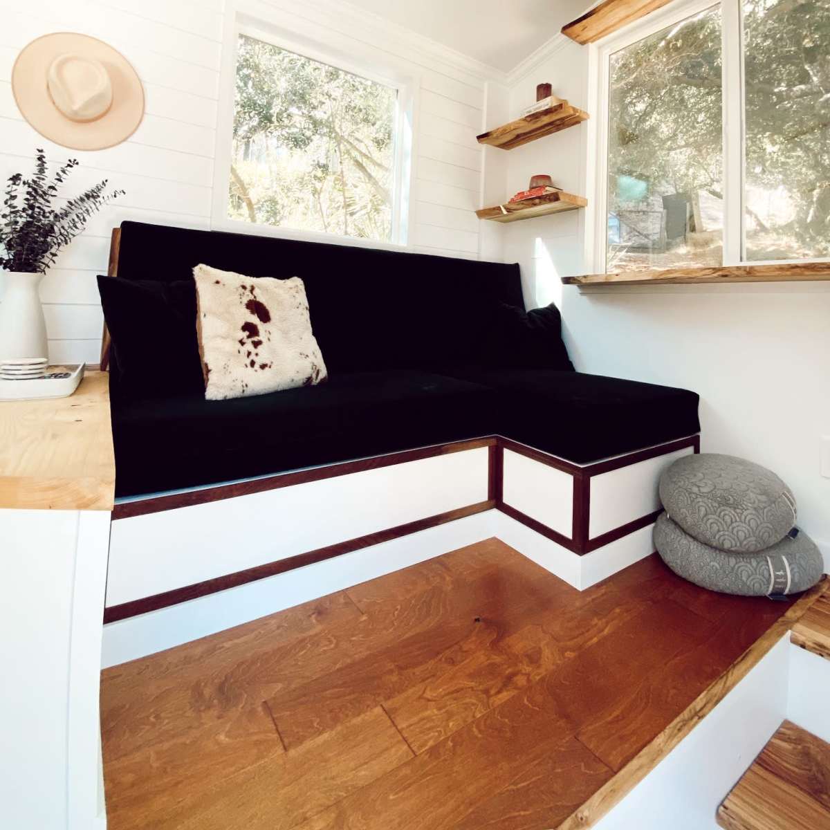 A cozy interior space with a black couch, wooden steps, and large windows, illuminated by natural light.
