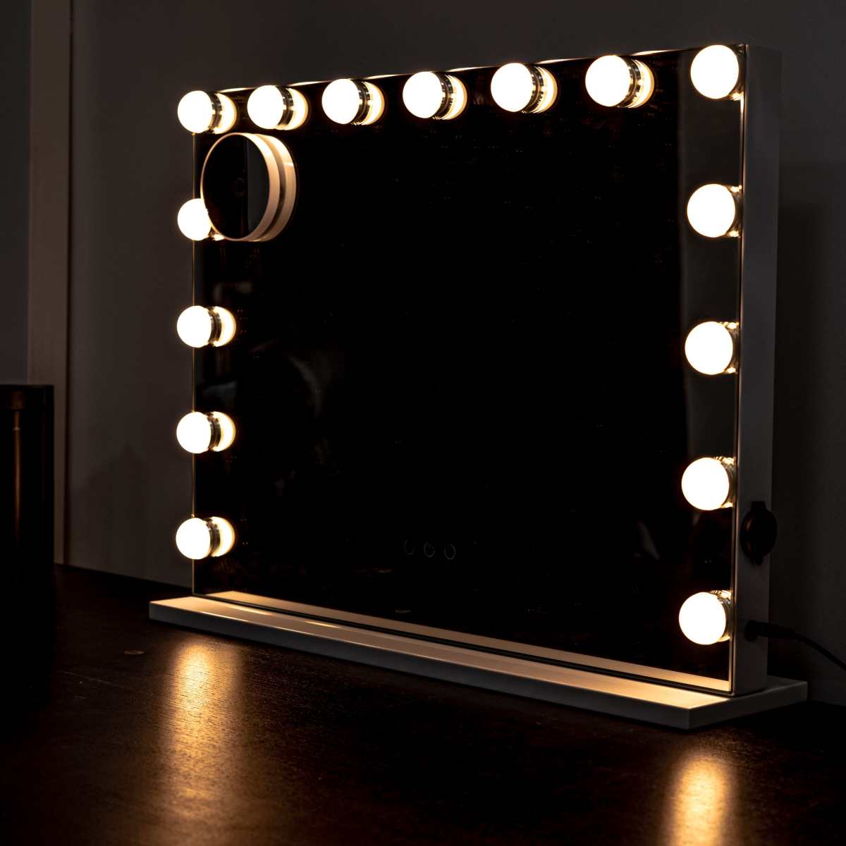 A rectangular makeup mirror with bright circular bulbs surrounds its frame is positioned on a dark surface next to a smaller illuminated mirror.