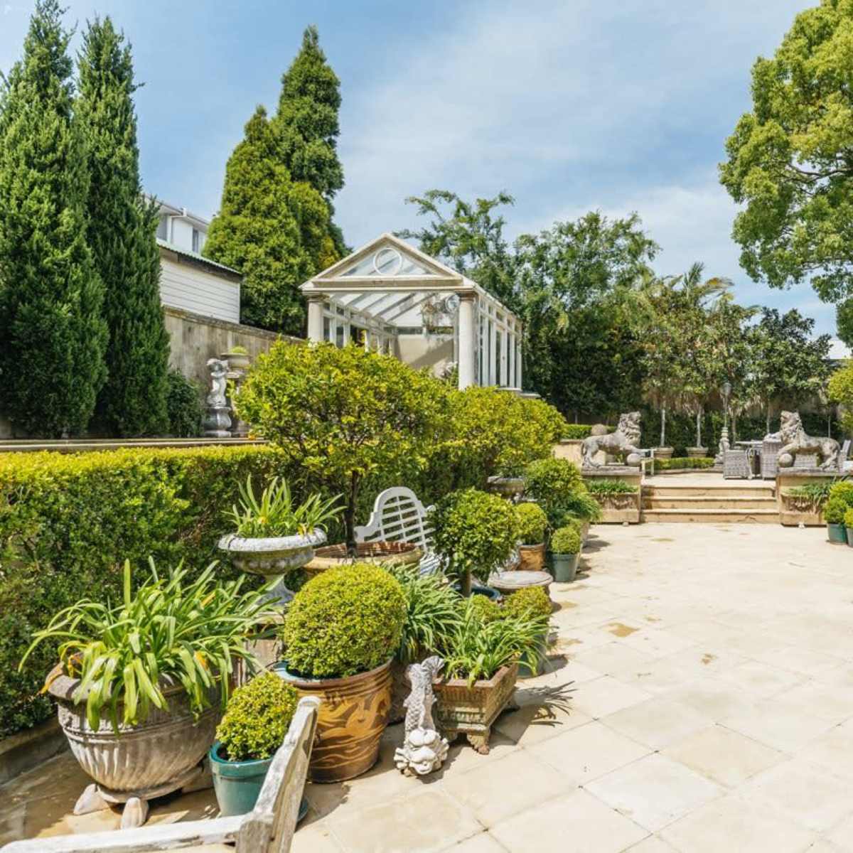 A landscaped outdoor garden area with stone pathways, manicured hedges, and potted plants arranged around ornamental sculptures.
