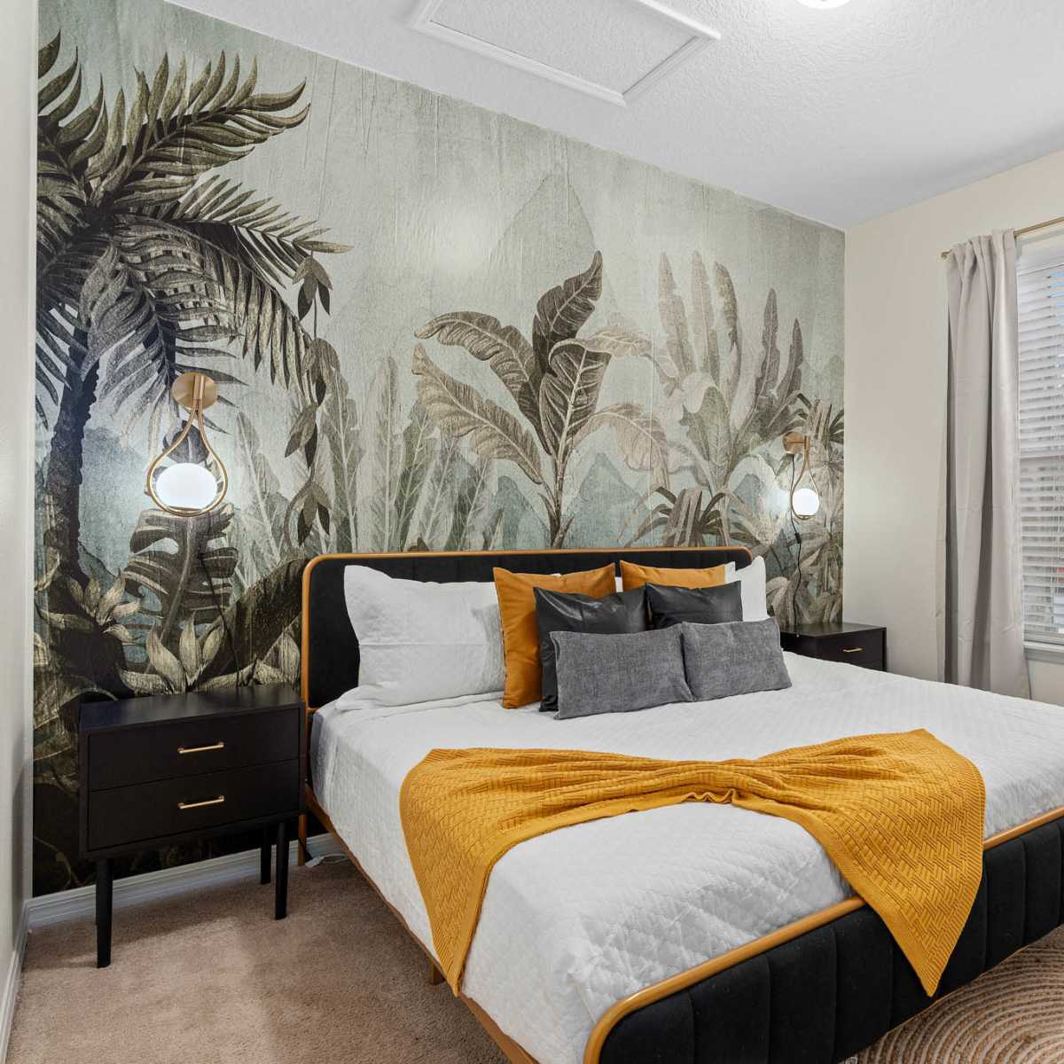 A bedroom features a large bed with an orange and gray throw blanket, flanked by two nightstands, and adorned with a tropical mural and potted plant.