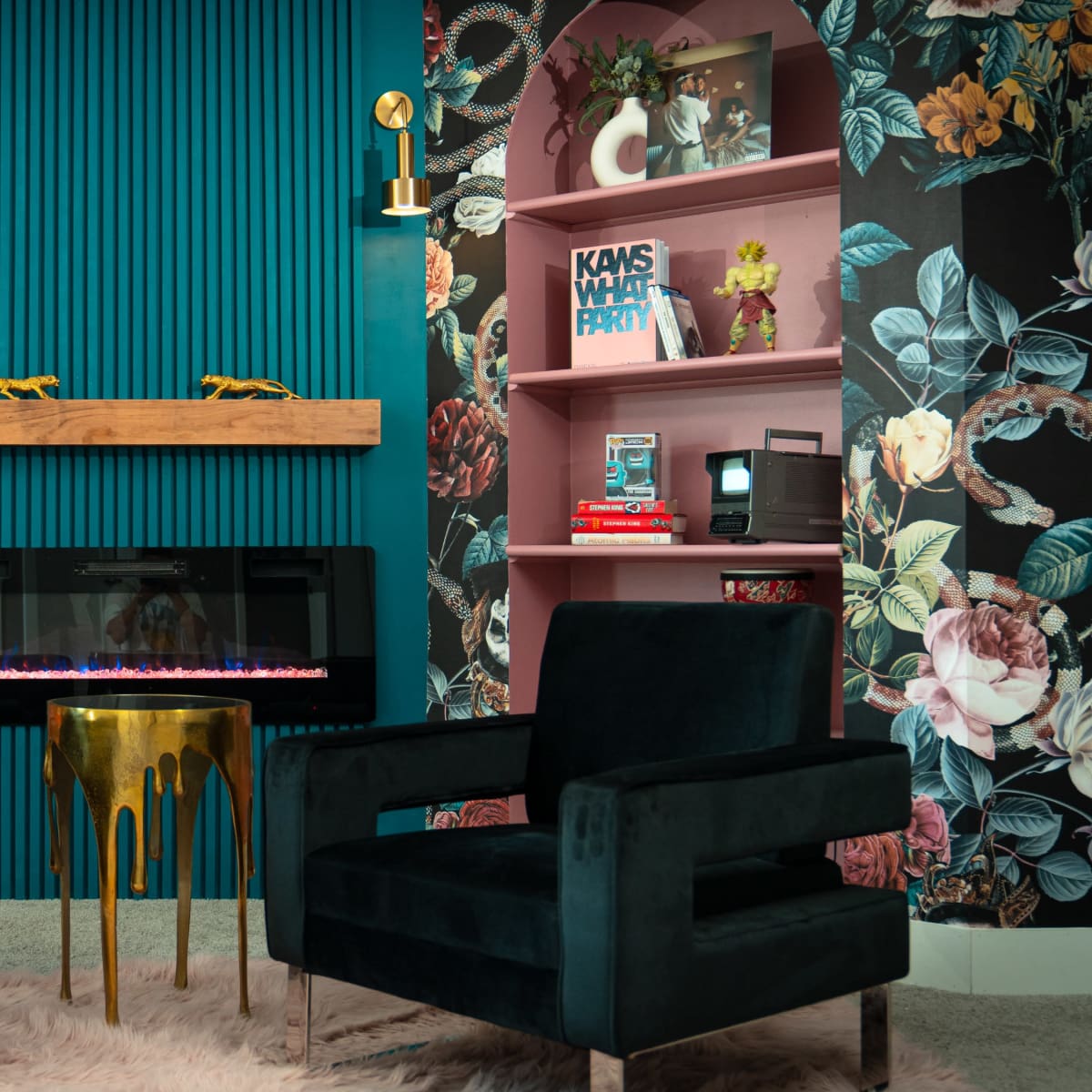 A cozy seating area with two dark green chairs facing a modern electric fireplace, surrounded by floral-patterned wallpaper and pink shelves displaying various decorative items.