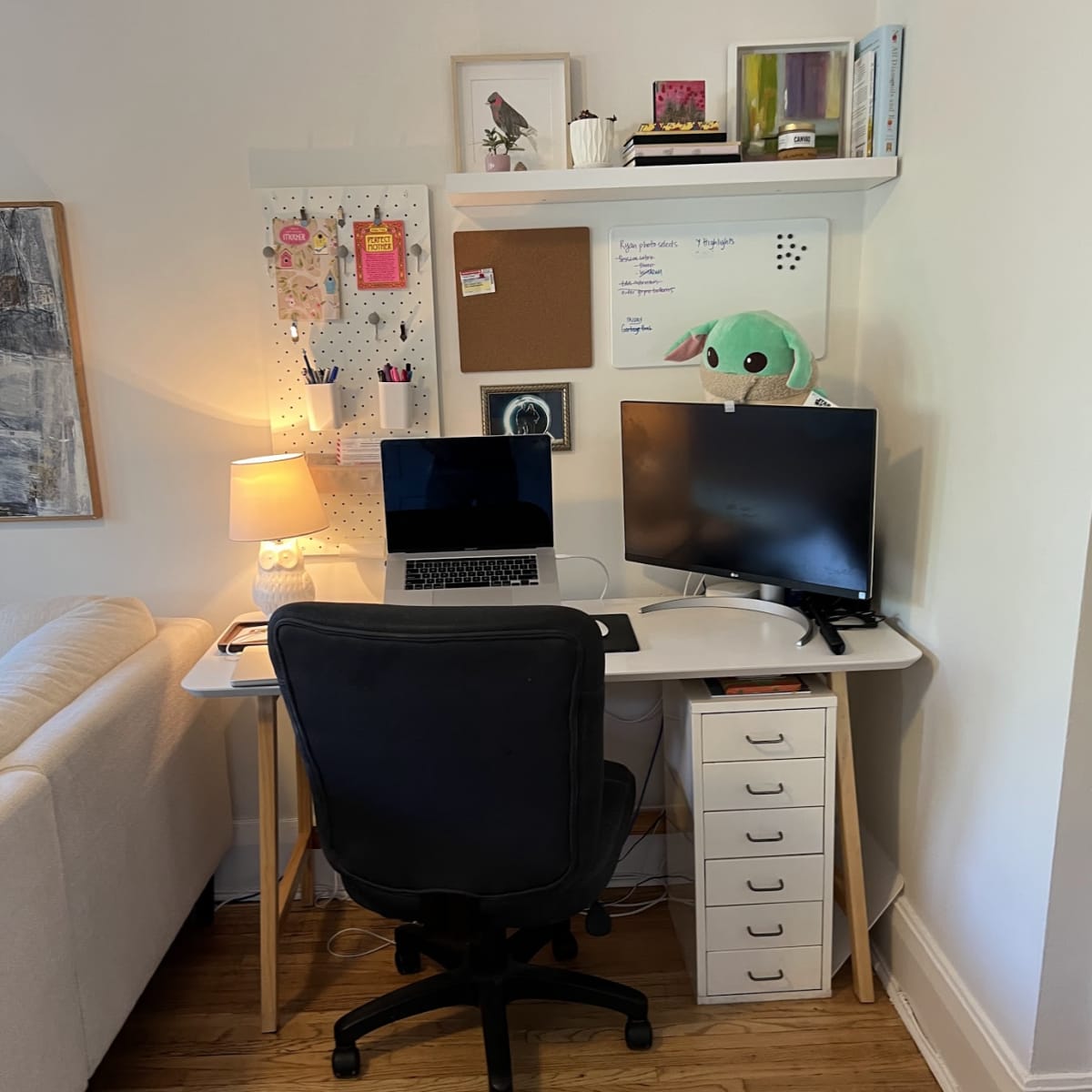A small home office setup features a desk with a laptop, a monitor, a lamp, and a cork board mounted on the wall, accompanied by a plush toy on the shelf above.