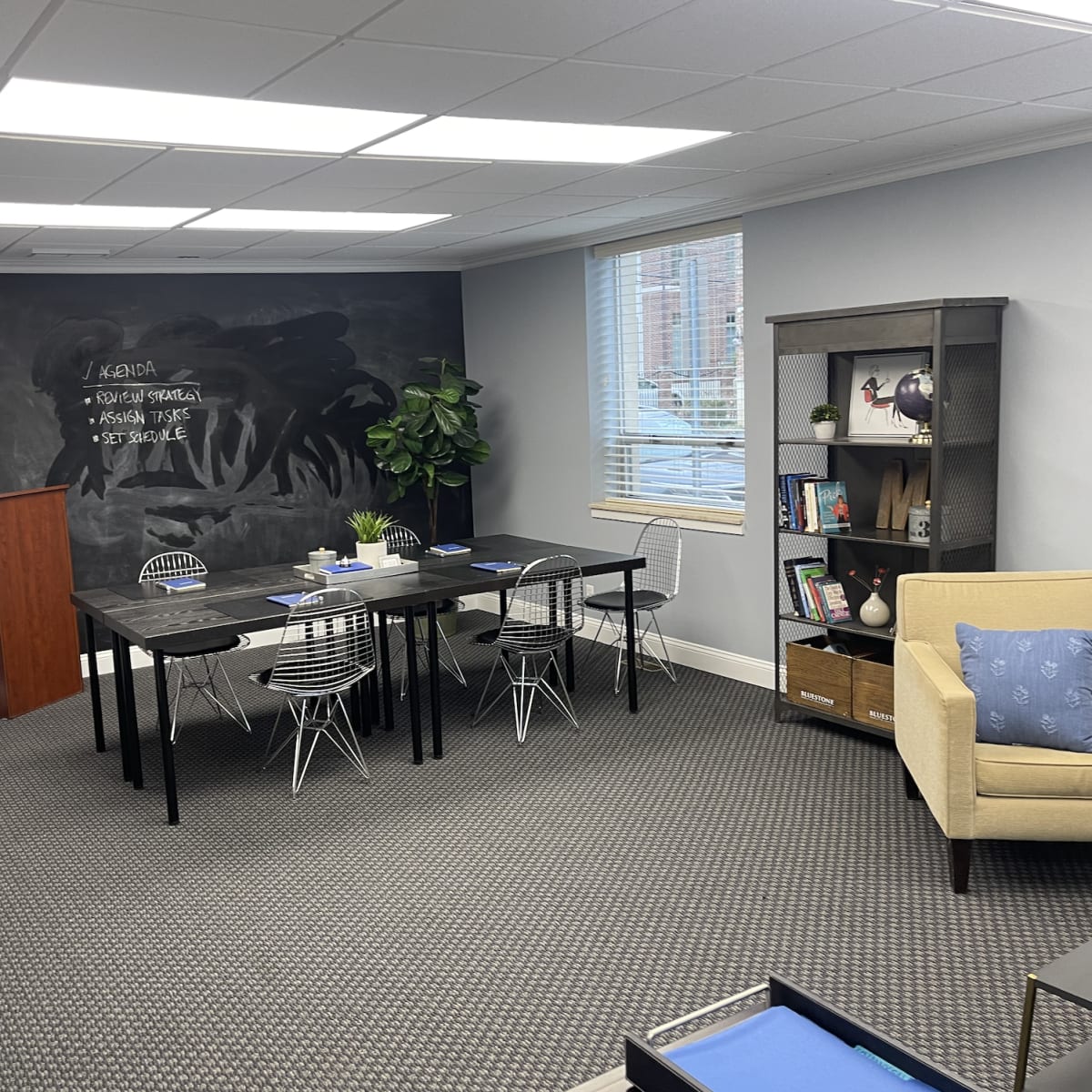An office space featuring a black chalkboard wall, a dining area with several black tables and white chairs, a small bookshelf, and a beige armchair in one corner.