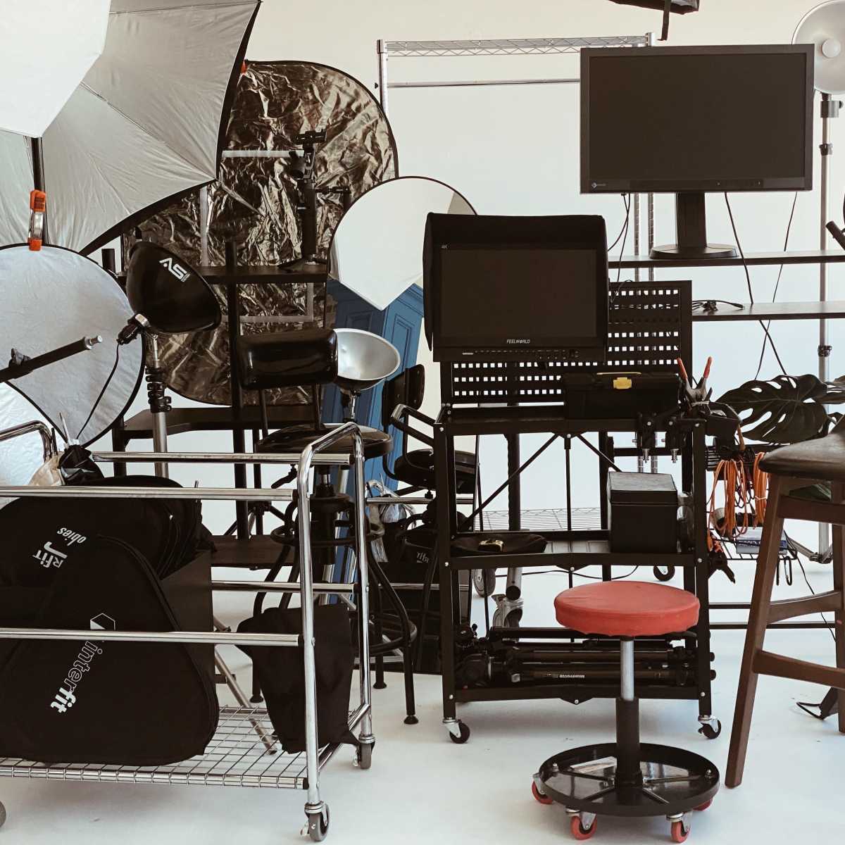 A photography studio setup with various lighting equipment, reflective umbrellas, a computer monitor, and storage carts.