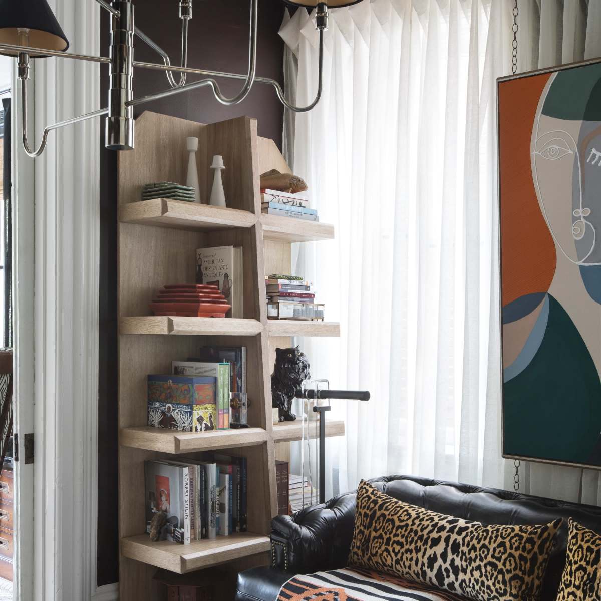 A black leather sofa with a patterned throw pillow is positioned next to a wooden bookshelf filled with books and decor, beneath a chandelier and large windows with sheer curtains.