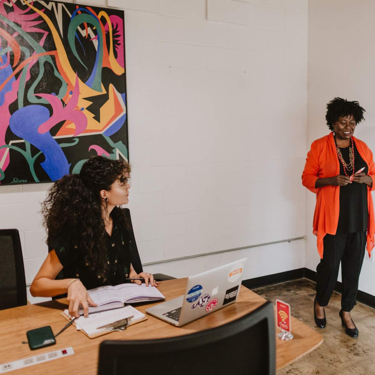 A woman in an orange blazer speaks to a seated woman at a table with a laptop, in a modern meeting room adorned with colorful artwork.