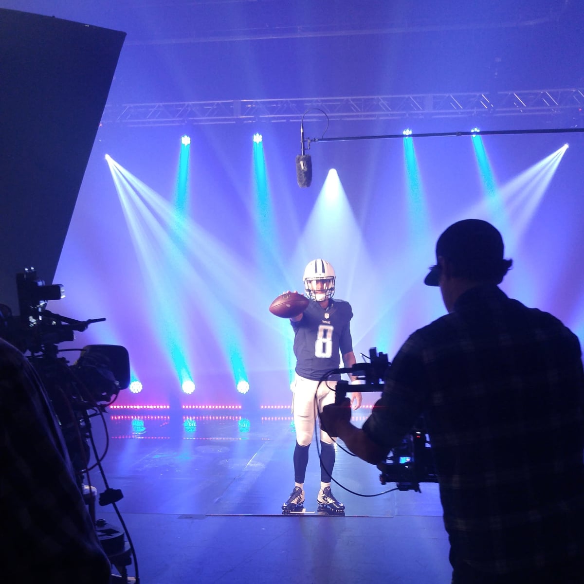 A football player poses in a studio under bright lights while a crew records the scene.