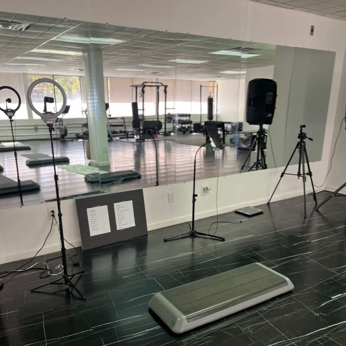 A fitness studio with a step platform in the center, surrounded by cameras and lighting equipment, and a mirror reflecting the space.