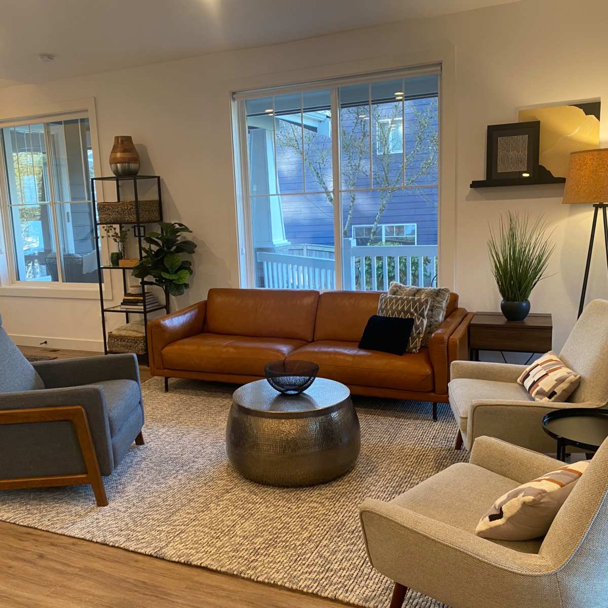 The living room features a brown leather sofa, two gray armchairs, a round metallic coffee table, and large windows showcasing outdoor greenery.