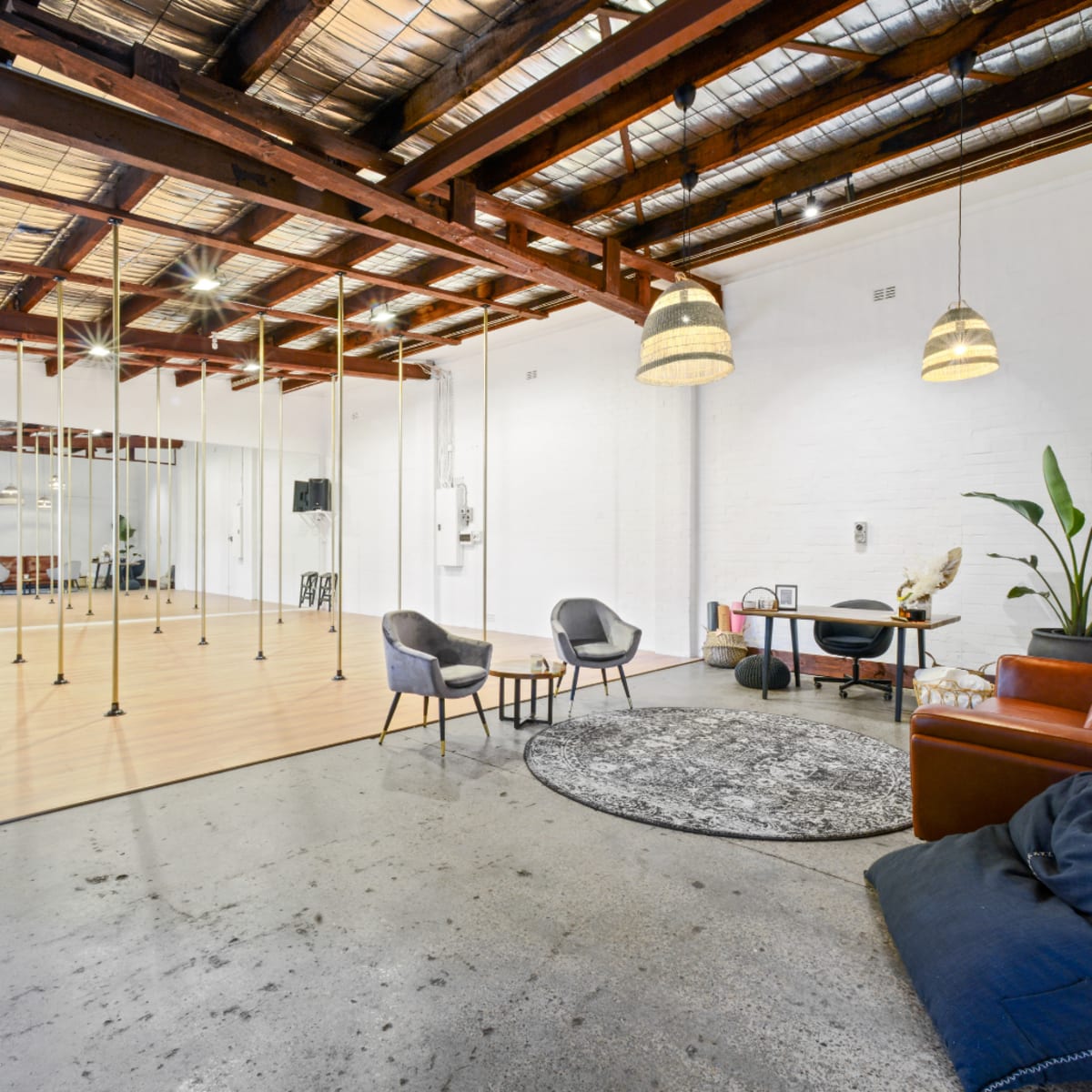A spacious studio with wooden beams, a mirrored wall, and a mix of seating areas, including chairs and rugs, on a polished concrete floor.