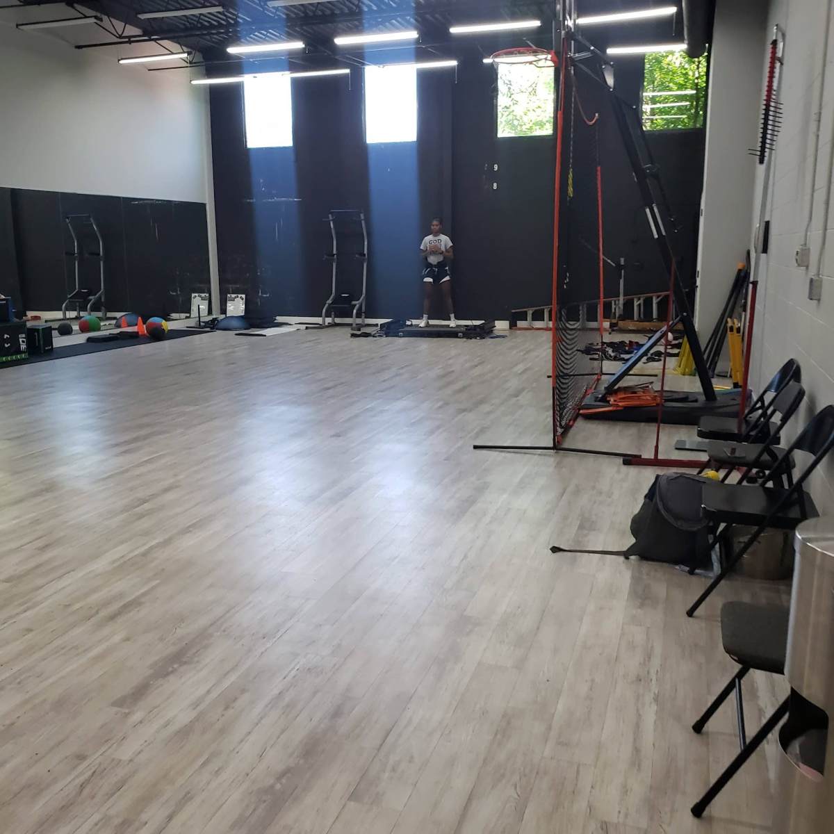 An indoor gym or training facility with equipment along the walls, a person standing at the back, and empty chairs arranged on one side.