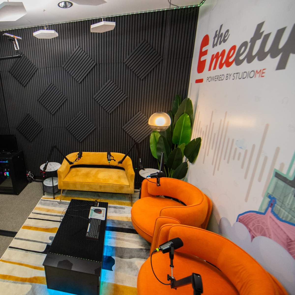 A modern recording studio with two orange chairs, a yellow couch, a striped rug, and a television mounted on the wall displaying the logo "the meetup."