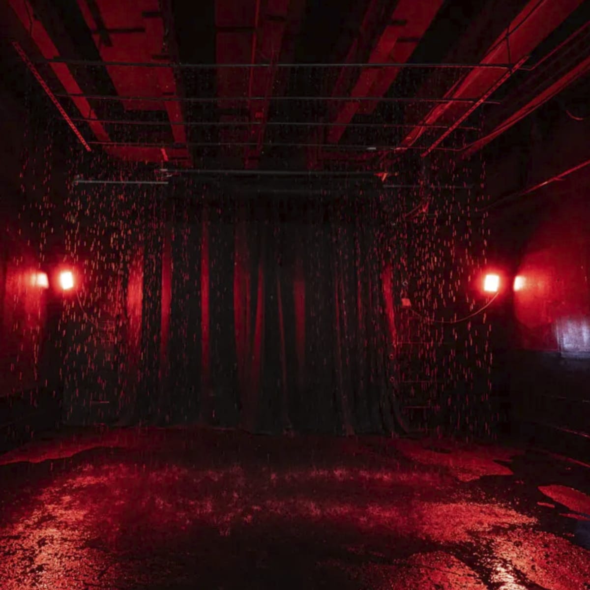 A dark room with red lighting, a black curtain in the background, and water cascading down from the ceiling onto the wet floor.