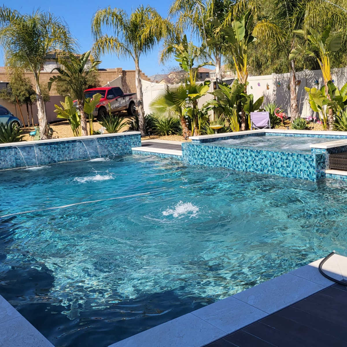 A swimming pool area featuring a main pool with a spa section, surrounded by palm trees and landscaped plants.