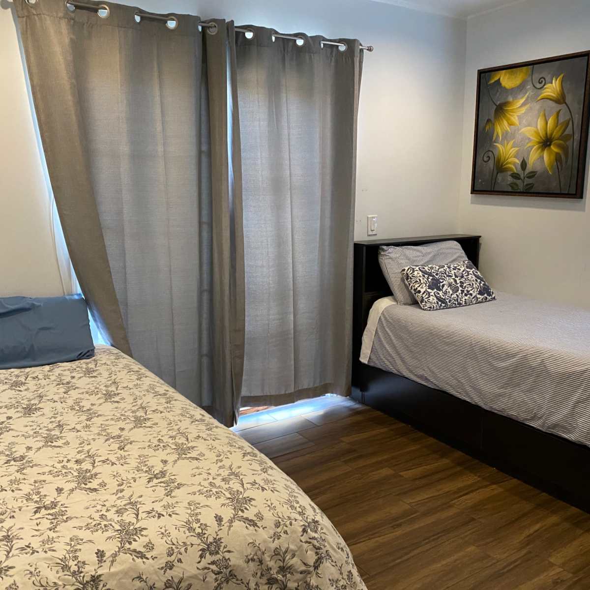 A bedroom with a double bed covered in a floral comforter beside a twin bed against a wall, with gray curtains and a painting of yellow flowers on the wall.