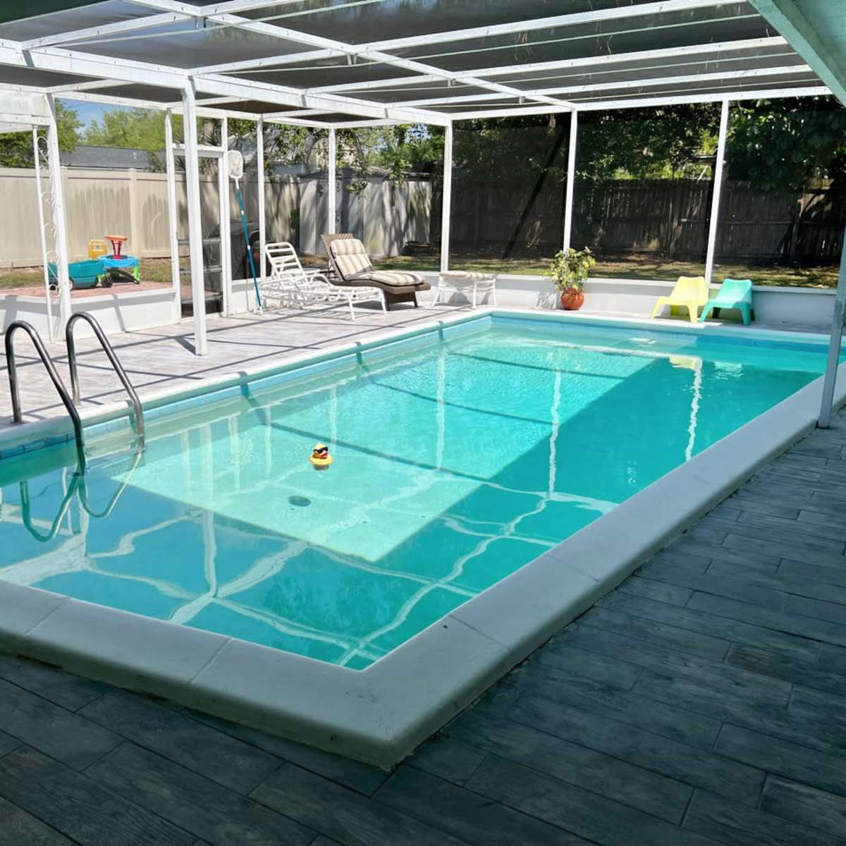 A clear swimming pool with a rubber duck floats in the water, surrounded by a screened patio area with lounge chairs and a children's play area in the background.