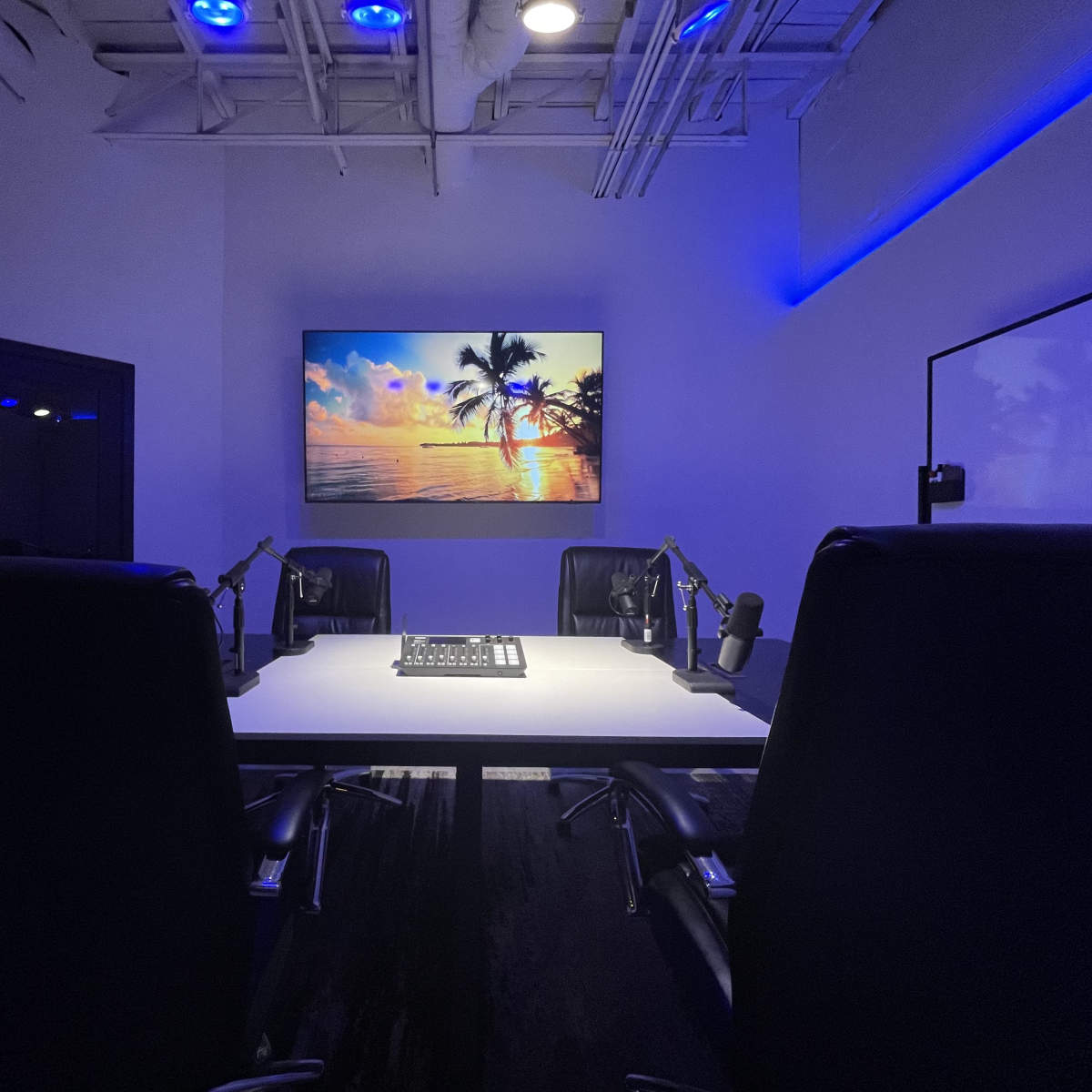 A dimly lit conference room features a large screen displaying a tropical sunset, with four black chairs arranged around a central table equipped with microphones.