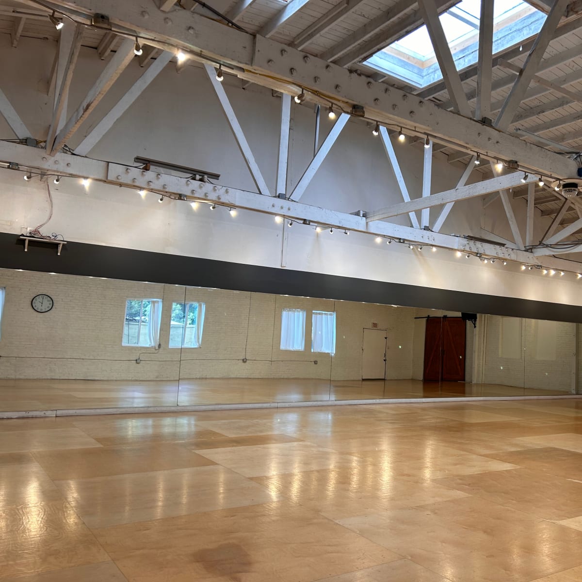 A spacious dance studio with wooden floors, large mirrors, exposed beams, and natural light coming through a skylight.