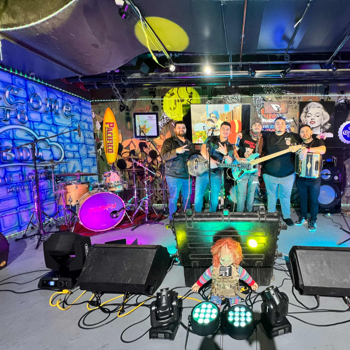 A group of five musicians poses on stage in front of a colorful, graffiti-covered backdrop with musical instruments and lighting equipment.