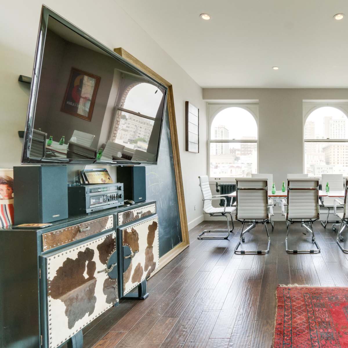 A modern office space with a long conference table surrounded by several chairs, a large television on the wall, and windows providing a view of a city skyline.