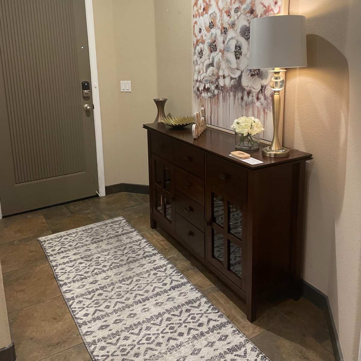 A small entryway features a dark wooden console table with decorative items and a table lamp against a light-colored wall, with a floral artwork hanging above and a green front door visible.