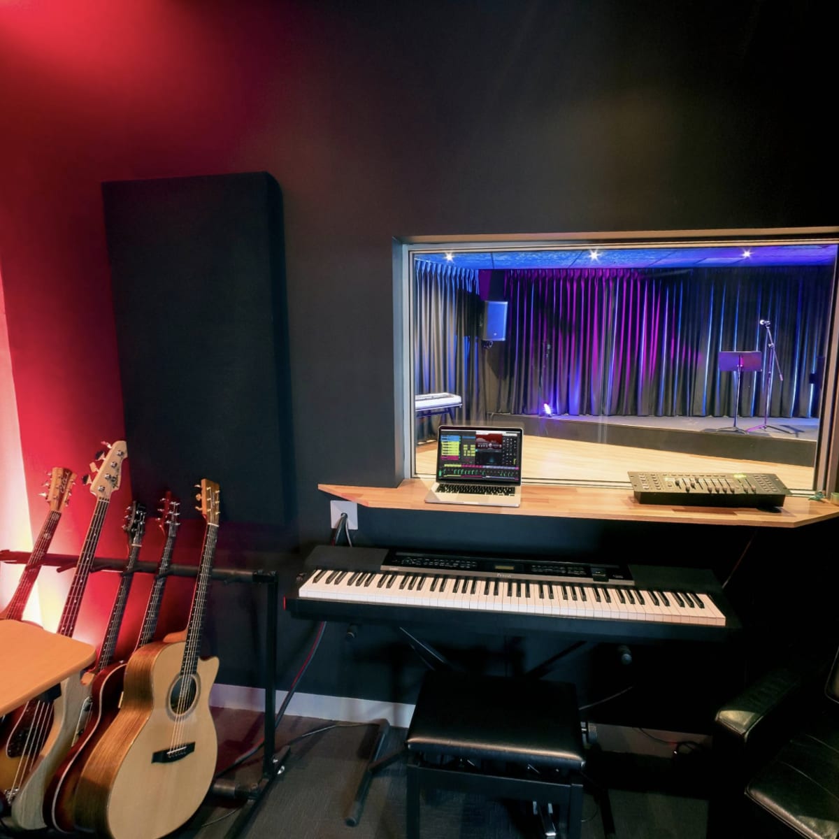 A recording studio featuring a piano, music production equipment, and guitars, with a window overlooking a stage area lit by purple and blue lights.