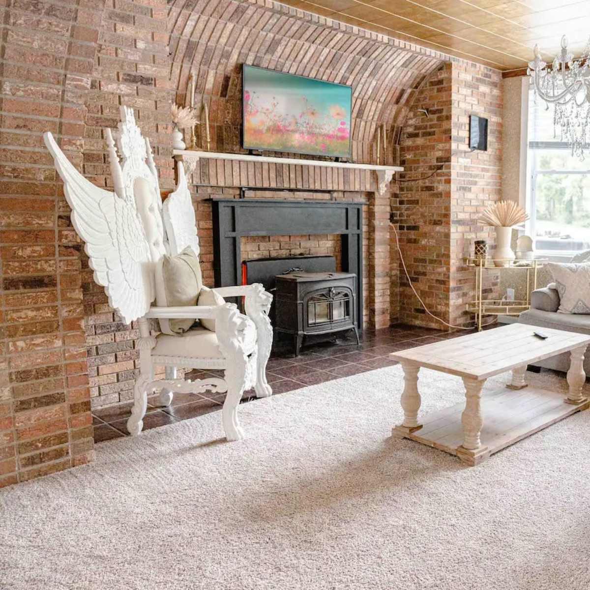 A cozy living room featuring a large sectional couch, a wooden coffee table, a brick fireplace with a TV above it, and a decorative chandelier.