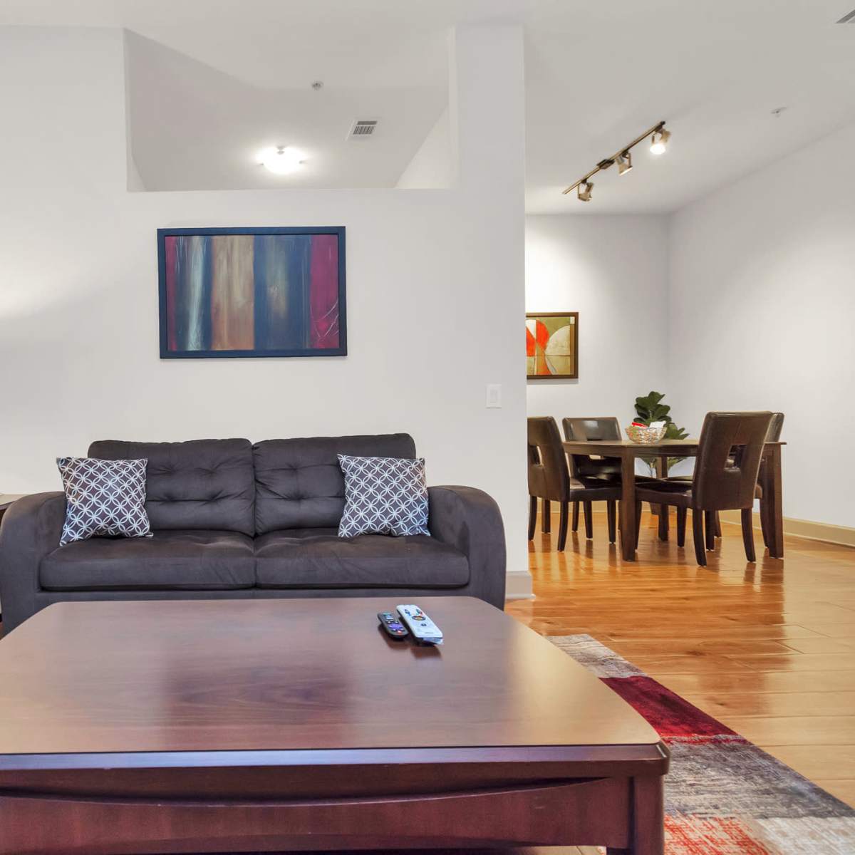 A living room features two dark sofas, a coffee table, and a lamp, with an adjacent dining area visible in the background.