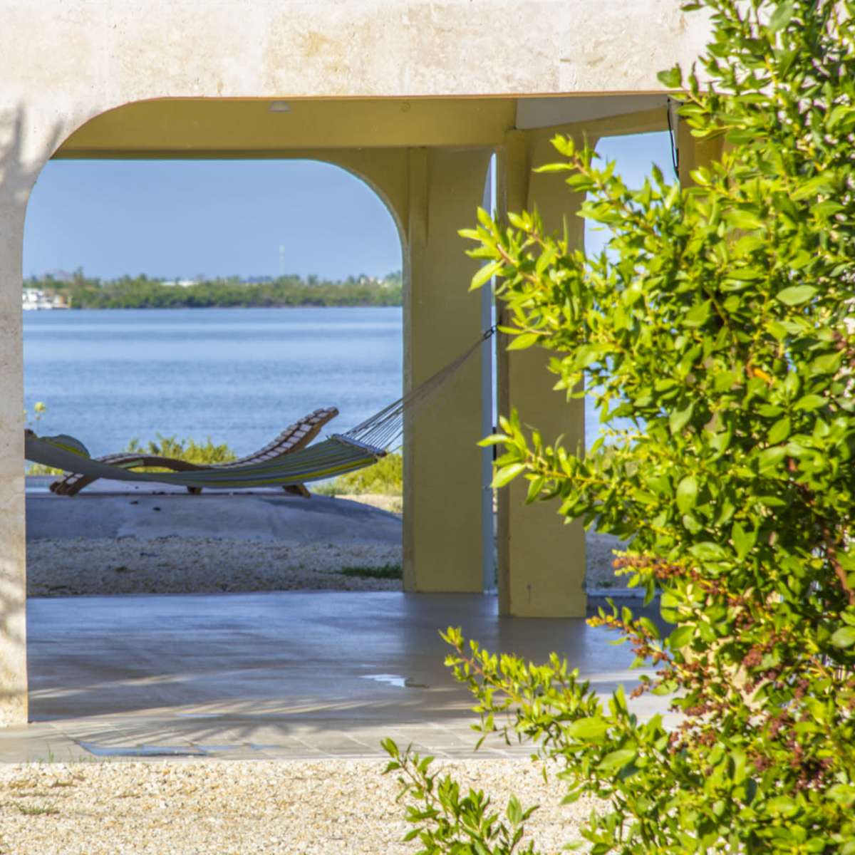 A hammock is suspended between two posts, framed by arches of a building, with a view of a calm body of water in the background.