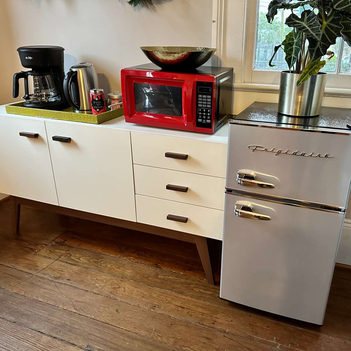 A compact kitchen sideboard features a coffee maker, a red microwave, and a silver refrigerator alongside potted plants.