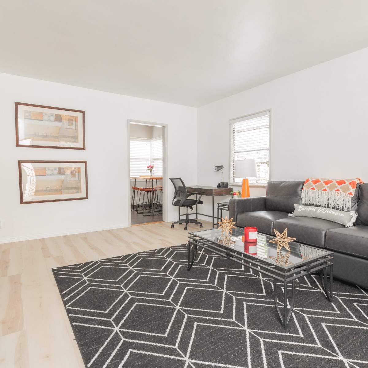 A living room with a dark sofa, a glass coffee table on a patterned rug, and two framed pictures on the wall.