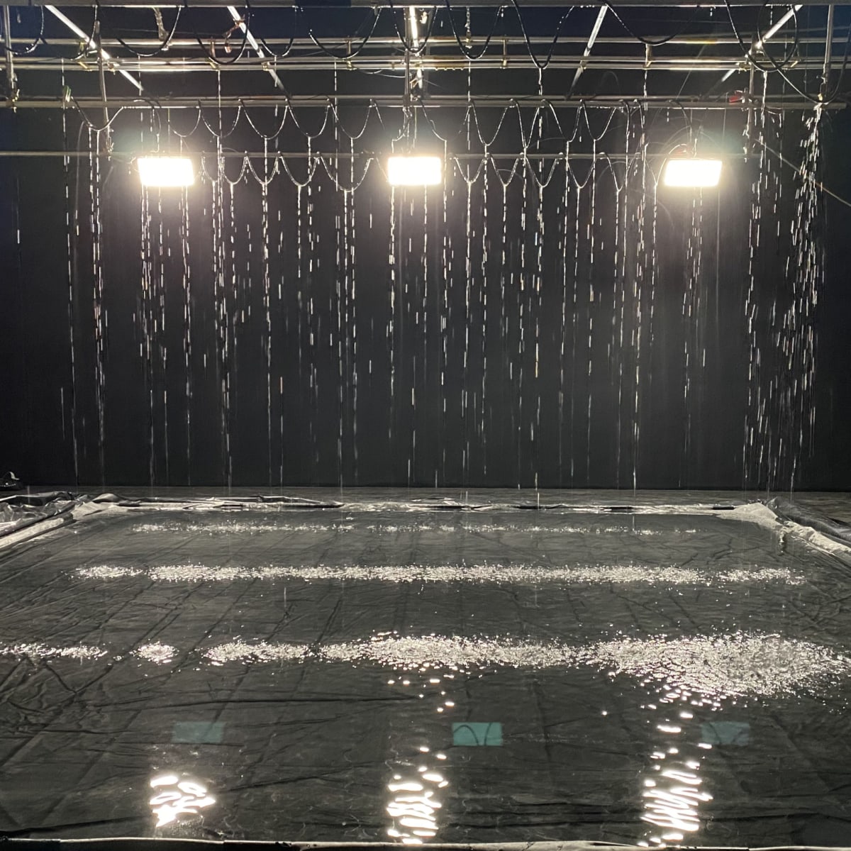 A black stage features a puddle of water with vertical streams of water falling from above under bright overhead lights.