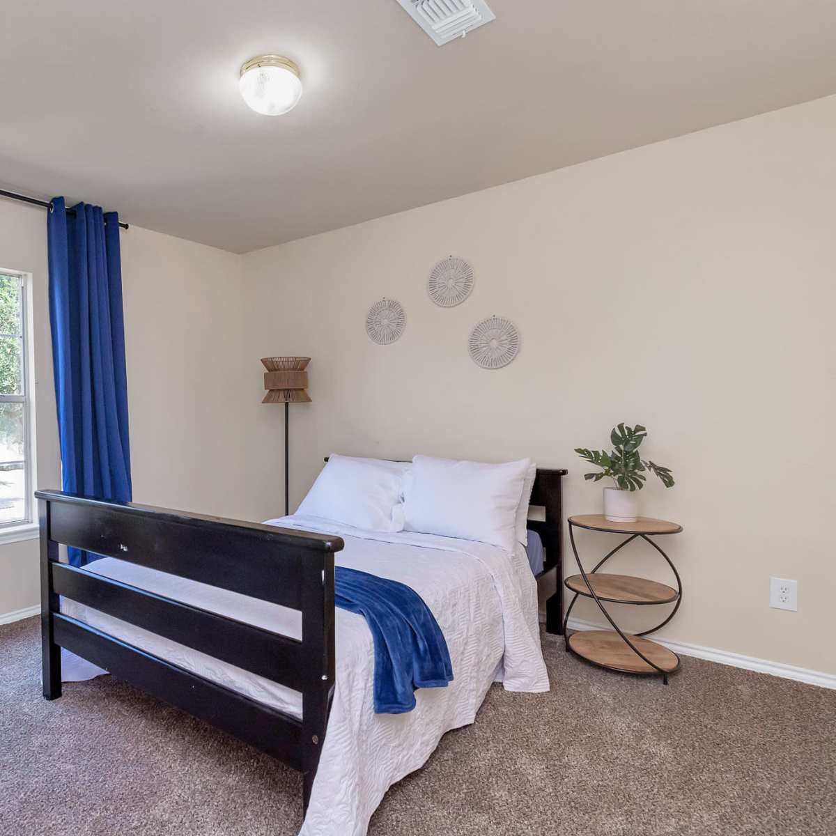 A simple bedroom features a bed with white linens, a round bedside table, and blue curtains framing a window.