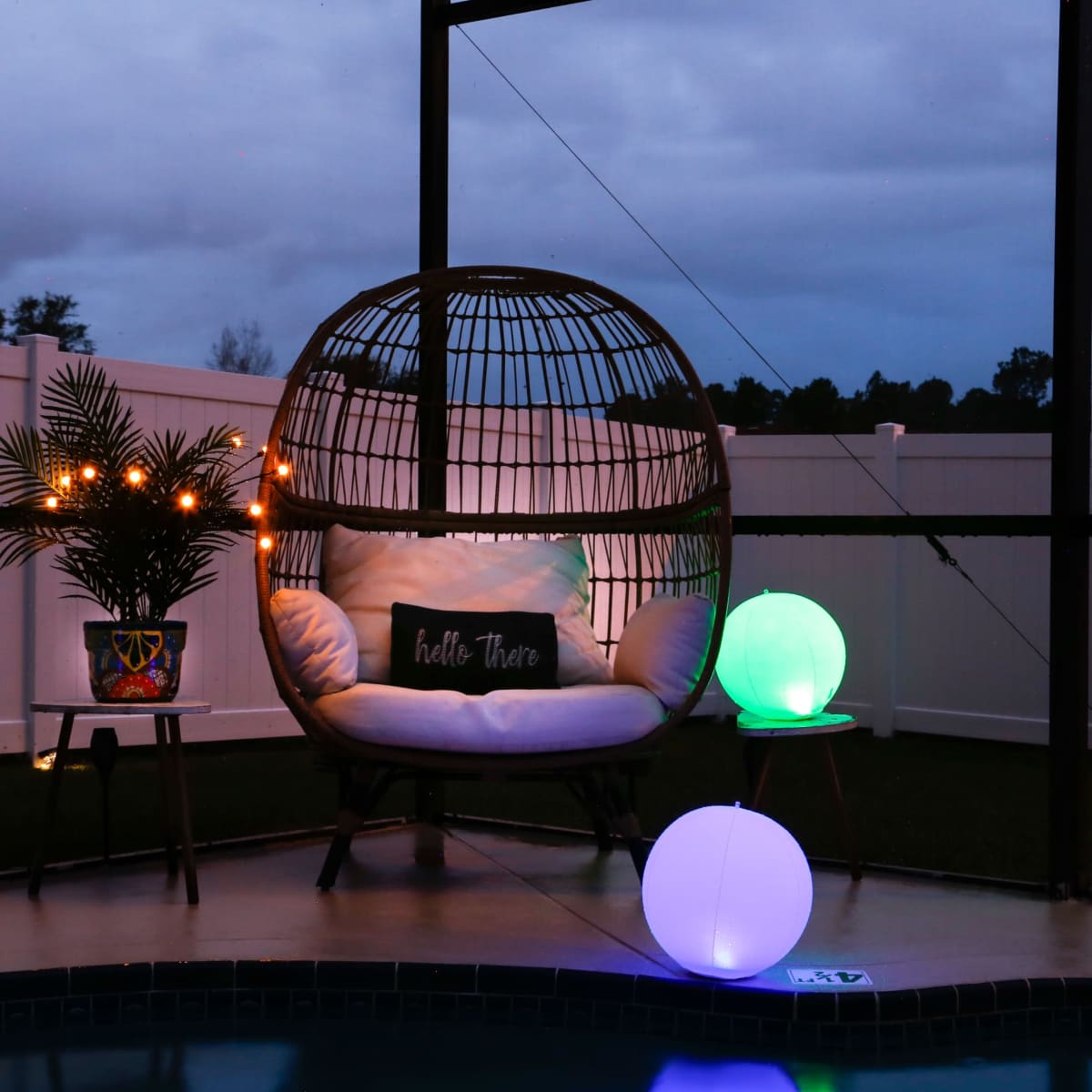 A hanging chair with cushions is situated beside a small table and illuminated orbs, set against a backdrop of a dimly lit outdoor area with a fence.