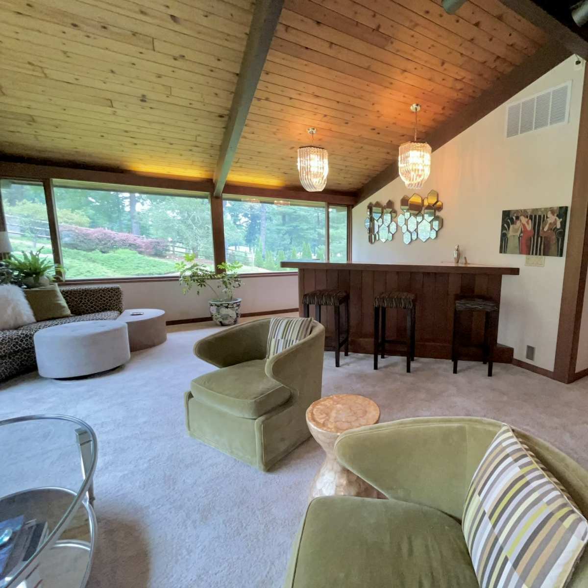A spacious living room with a bar area, featuring a wood-paneled ceiling and large windows overlooking greenery.