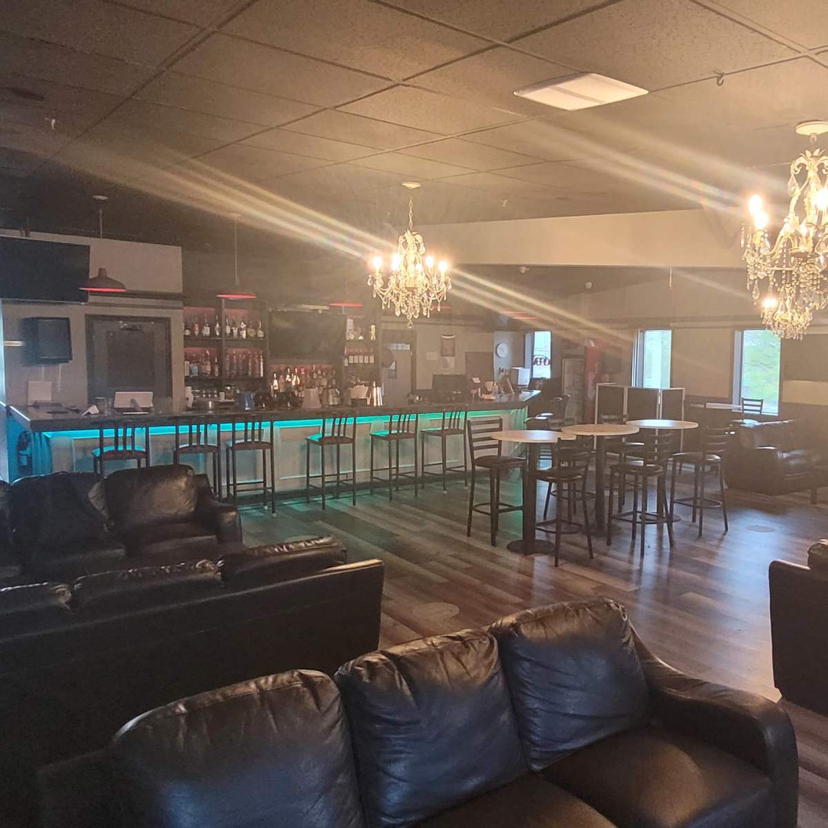 A spacious lounge area with black leather sofas, a bar with blue lighting, and chandeliers hanging from the ceiling.