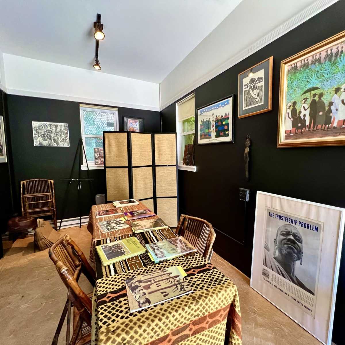 A room with black walls, a table covered with printed materials, and framed artwork displayed on the walls.
