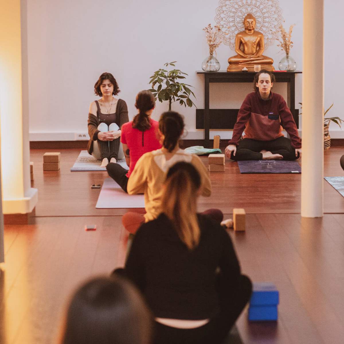 A group of people sits on yoga mats in a peaceful studio, facing a statue of a Buddha surrounded by soft lighting and plants.