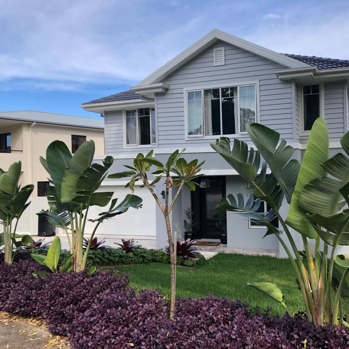 A two-story gray house with large windows is surrounded by tropical plants and a well-maintained garden.