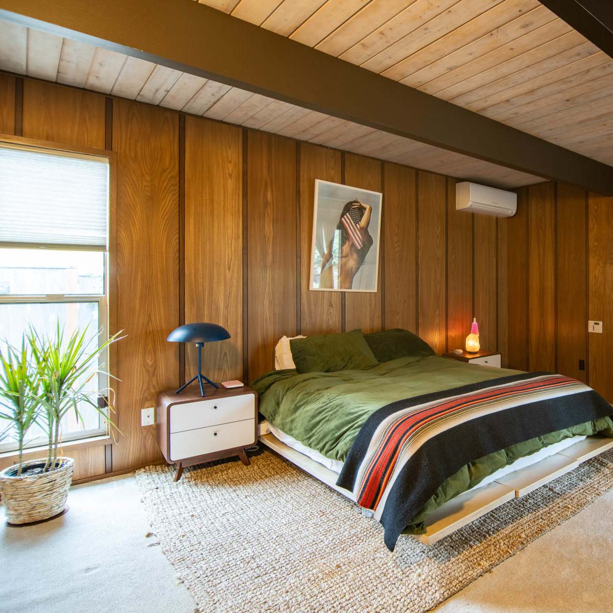 A wooden-paneled bedroom with a green bedspread, a lamp, a plant by the window, and artwork featuring a buffalo on the wall.