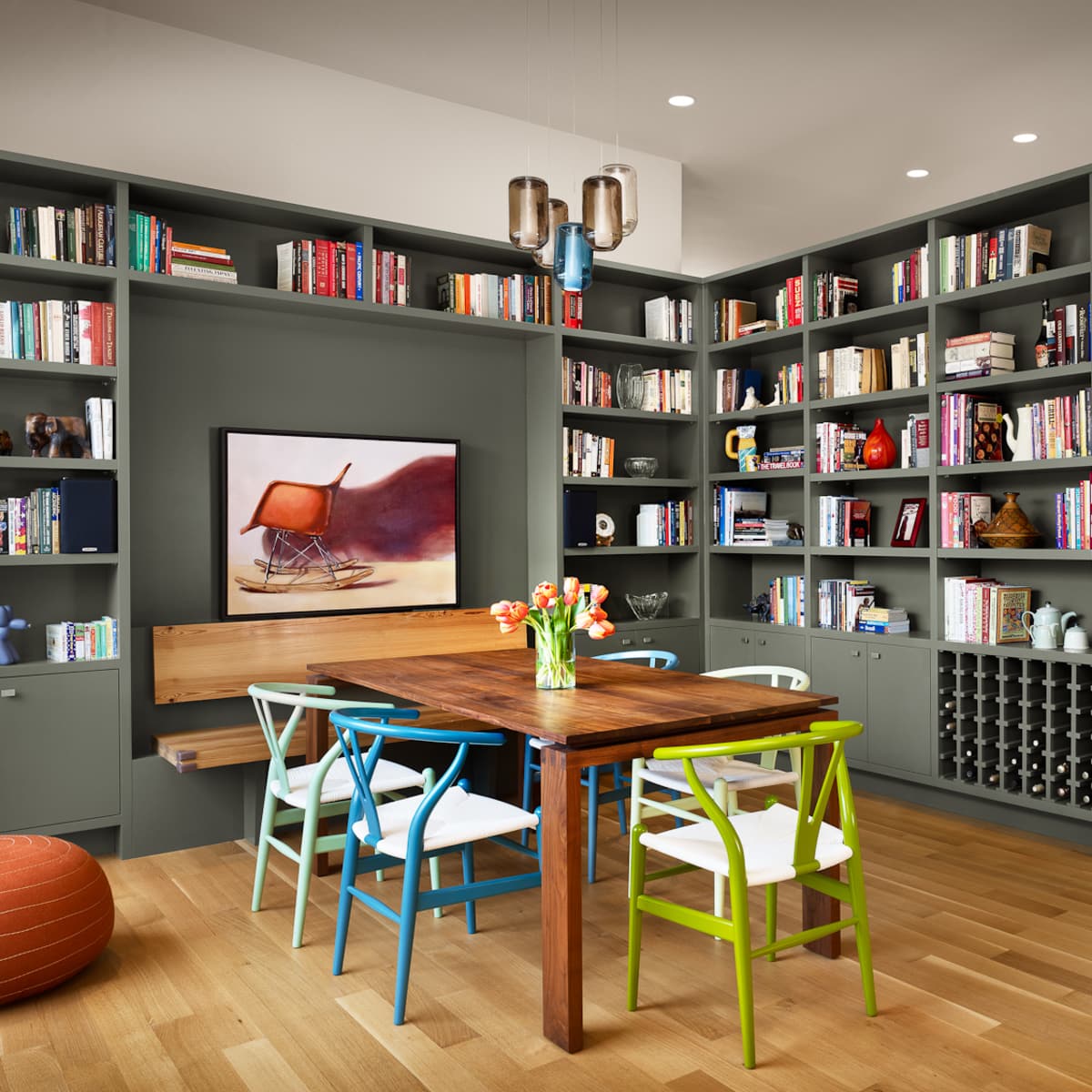 A modern dining area features a wooden table surrounded by colorful chairs, next to a bookshelf filled with books and decorative items.